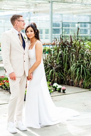 Wedding couple in white attire posing together in greenhouse with tropical plants.
