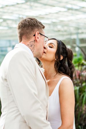 A romantic wedding kiss between a couple in white attire against a greenhouse backdrop.