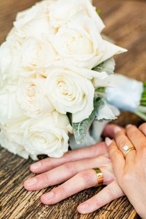 Close-up of hands wearing wedding rings next to white roses on a rustic wooden surface.