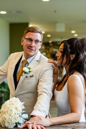 A romantic wedding couple poses together in white attire with cream roses at an indoor venue.