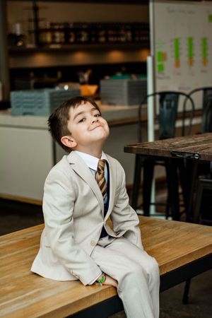 A young boy in a light-colored suit reclines on a wooden bench with a playful expression.