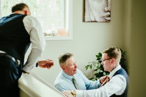 Pre-wedding preparations showing men helping with formal attire adjustments near a window.