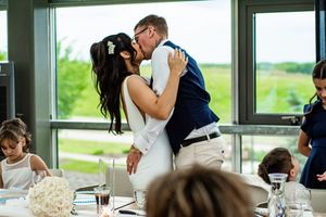 Wedding reception guests seated at long tables in a bright room with large windows overlooking green scenery.
