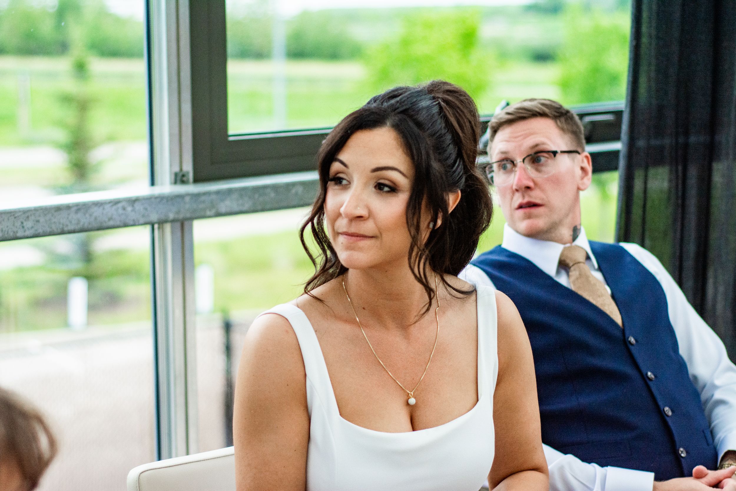Wedding guests in formal attire seated at tables during a reception in a bright venue with large windows.