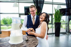 Newlyweds share a joyful moment while cutting their elegant white wedding cake by a window.