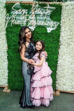 A woman in a dark dress poses with a young girl in a pink ruffled dress against a floral backdrop.