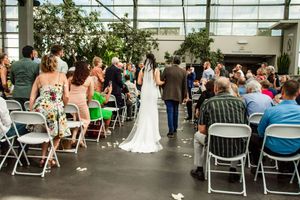 Wedding ceremony setup in a greenhouse with rows of white chairs and guests seated for the occasion.