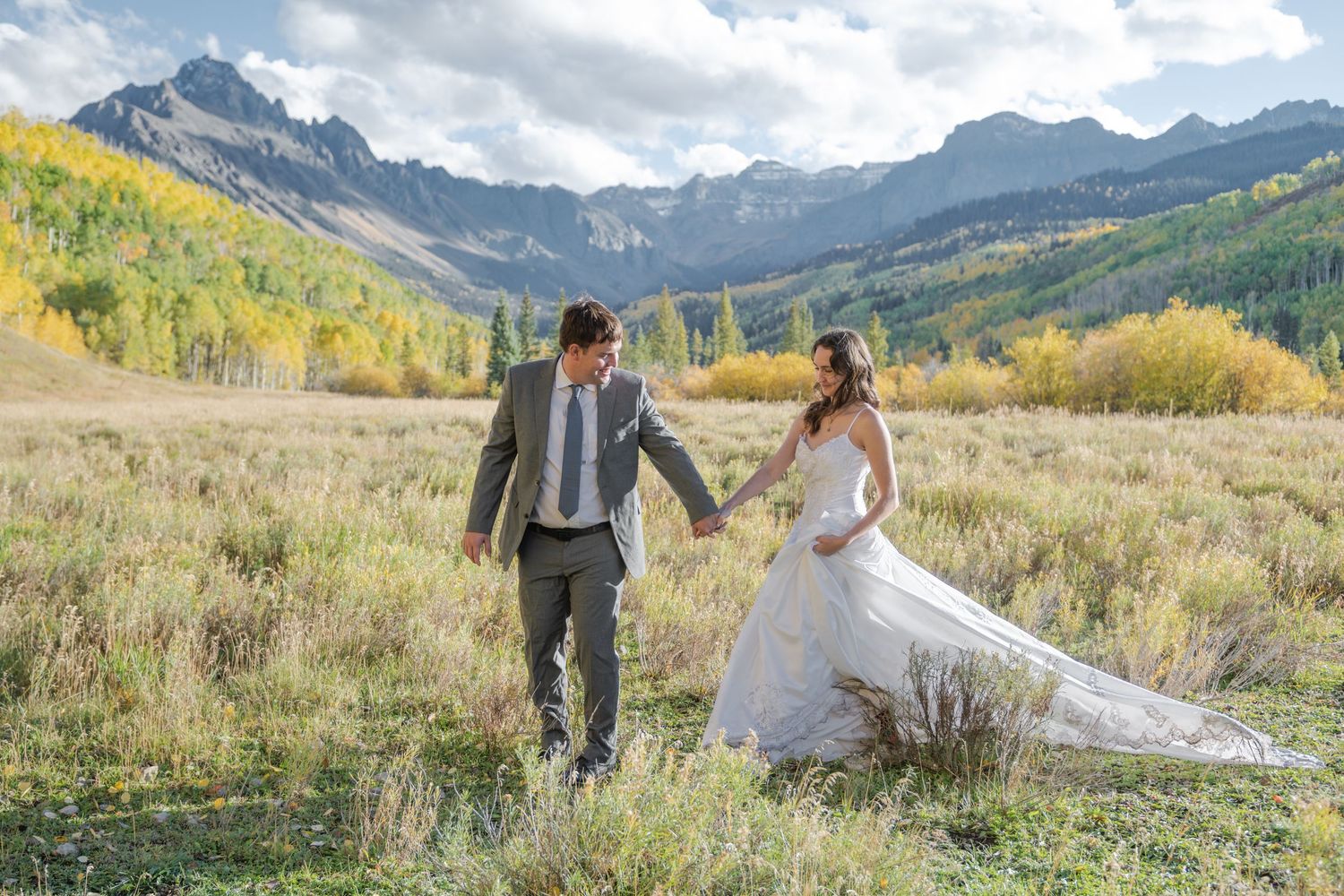 A bride and groom hold hands walking through a mountain meadow with autumn foliage and rocky peaks in the San Juan Mountains.