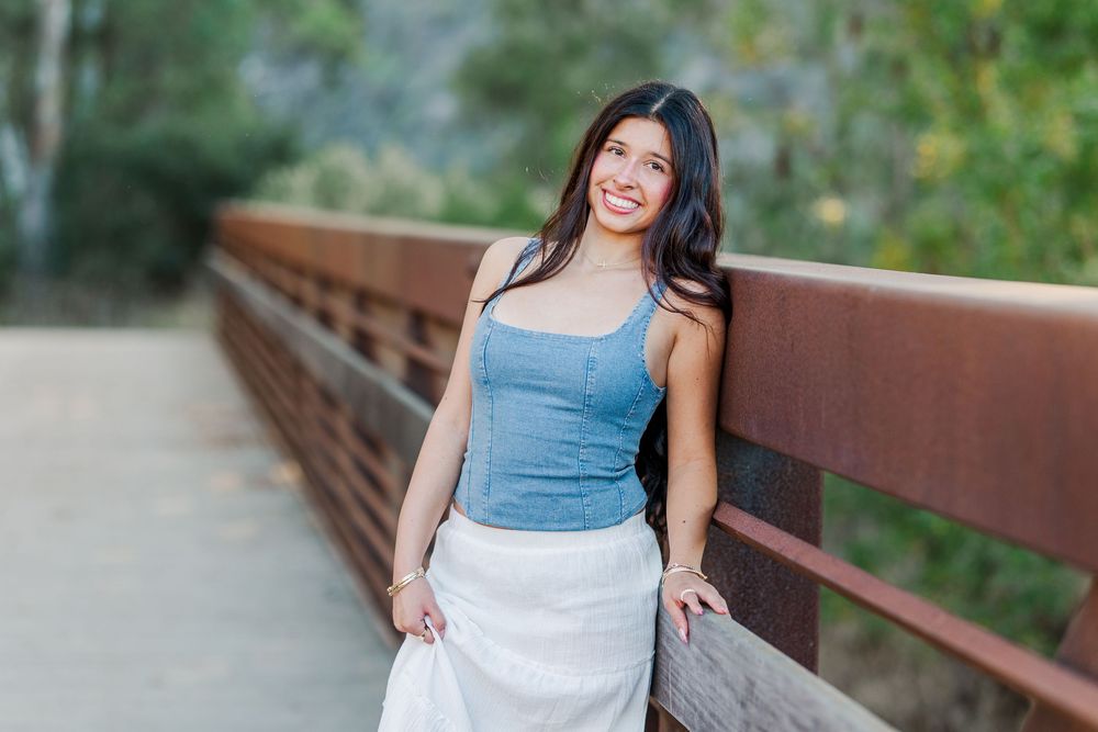 Woman in blue denim top and white skirt poses on rustic wooden bridge.