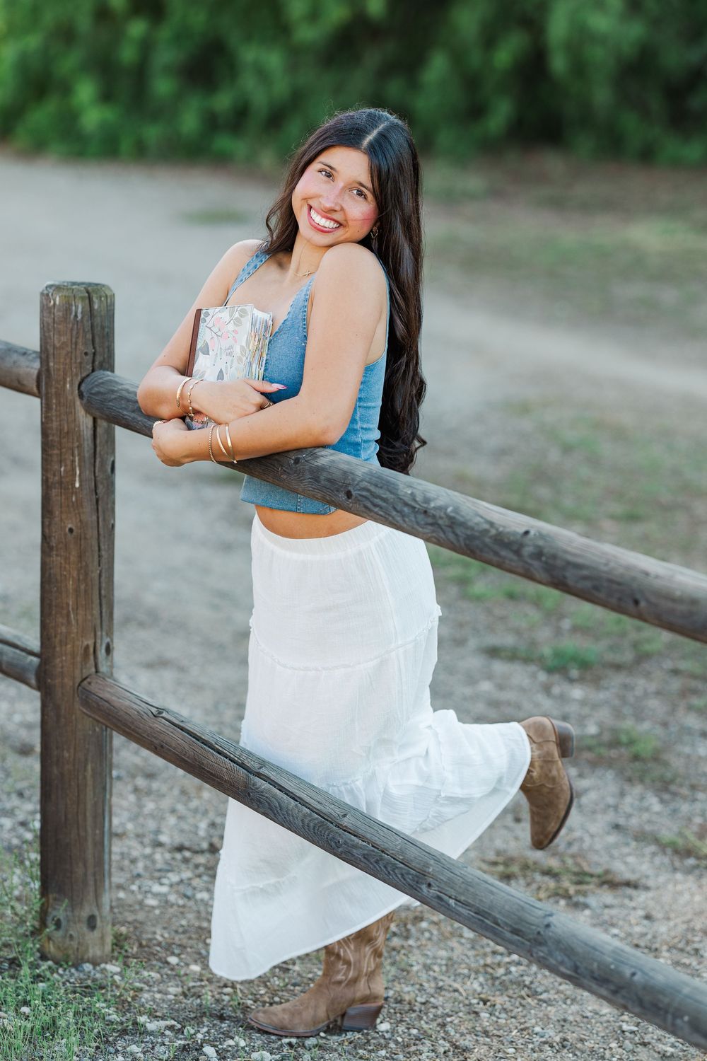 Woman in a white dress poses playfully by a rustic wooden fence in a natural outdoor setting.