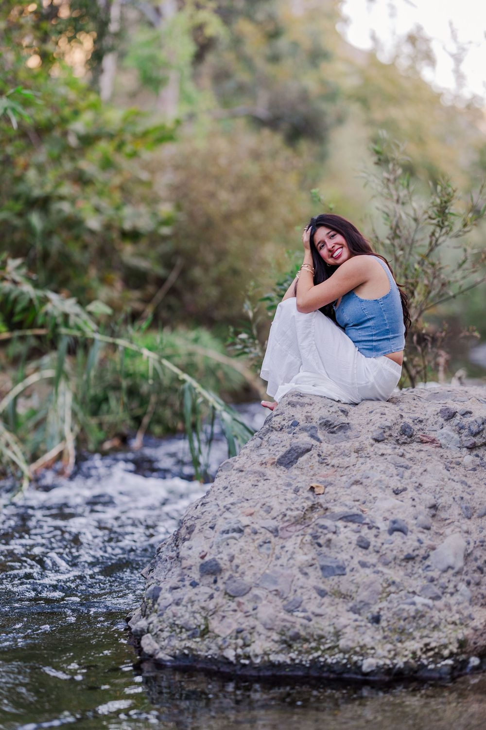 Woman relaxes on large rock by flowing stream in natural setting.