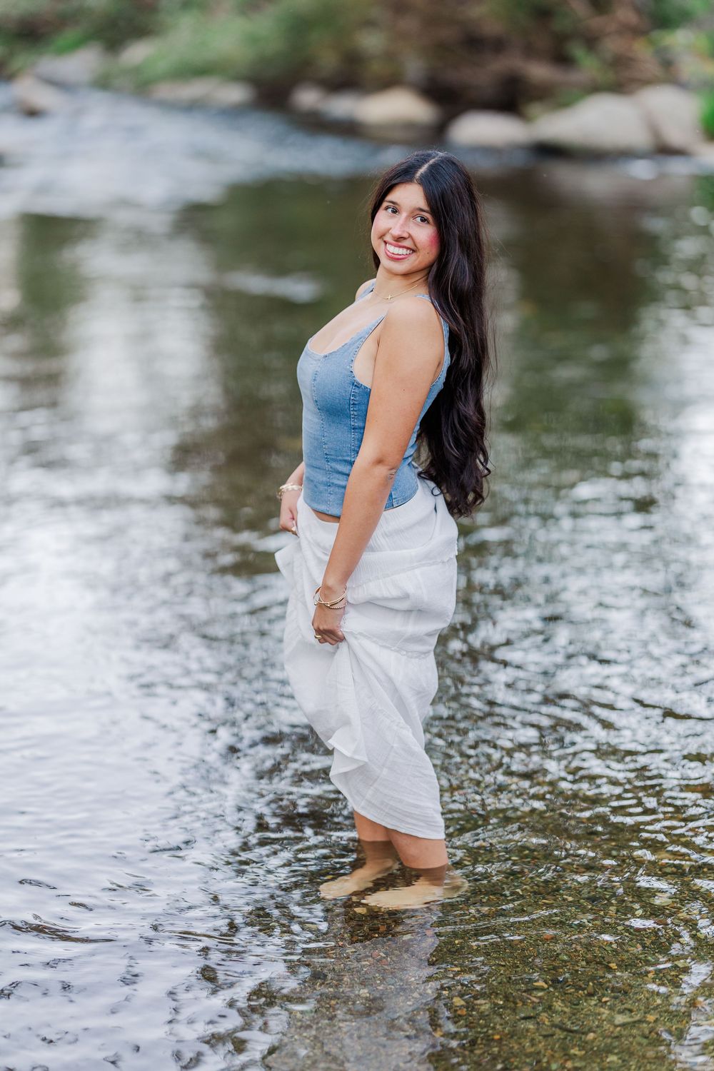 Woman in white dress and denim top wades through shallow flowing water during a sunny outdoor photoshoot.