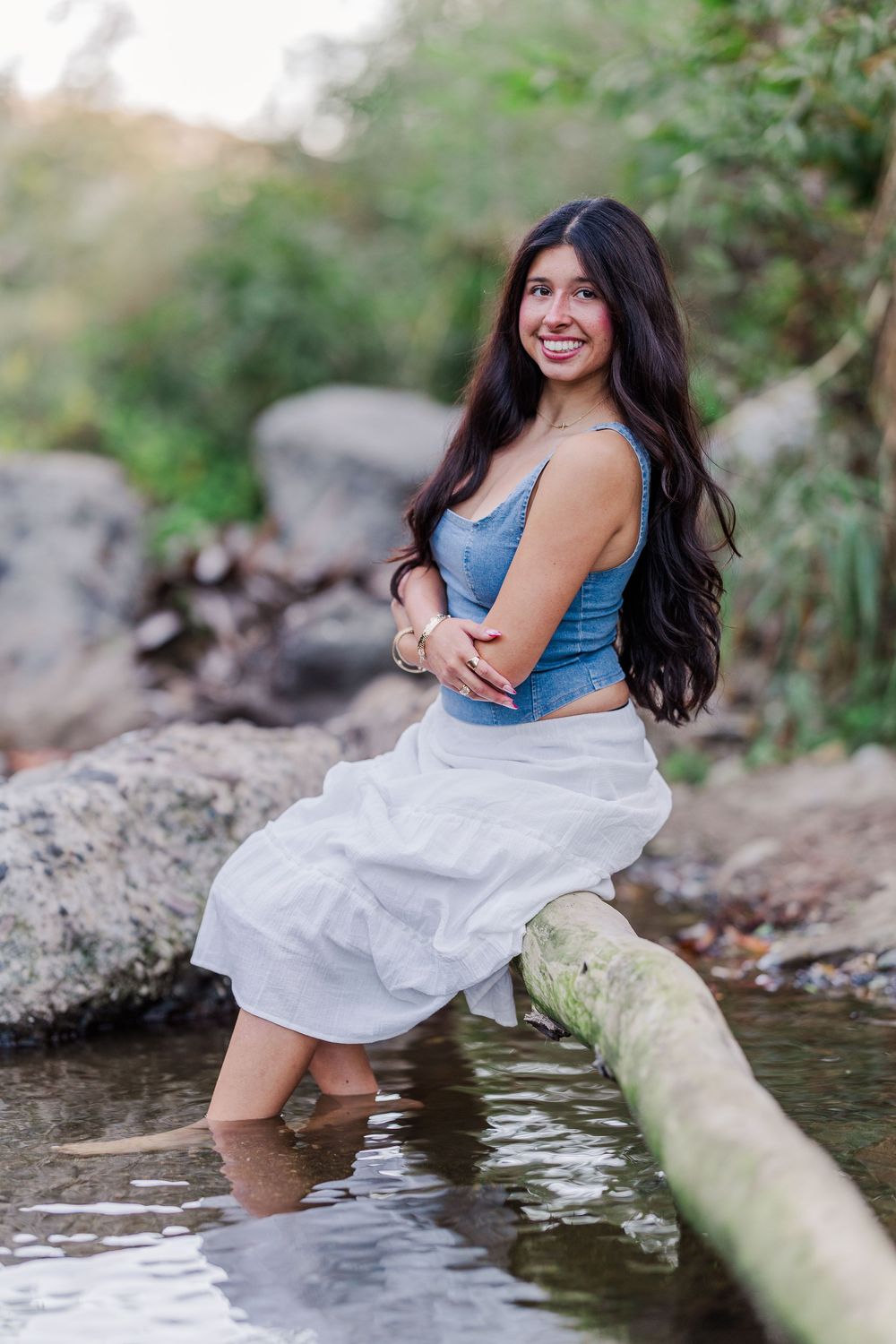 Playful poses by a stream wearing a white dress and denim top while sitting on rocks.