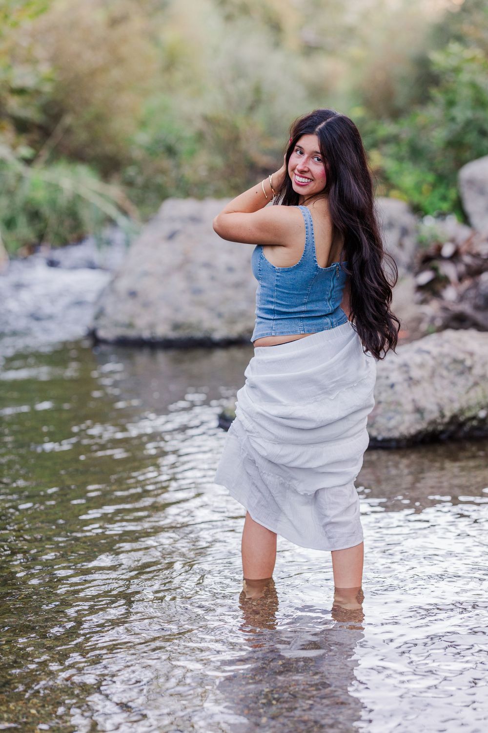 A woman in a blue top and white skirt poses playfully in a flowing stream surrounded by rocks and nature.