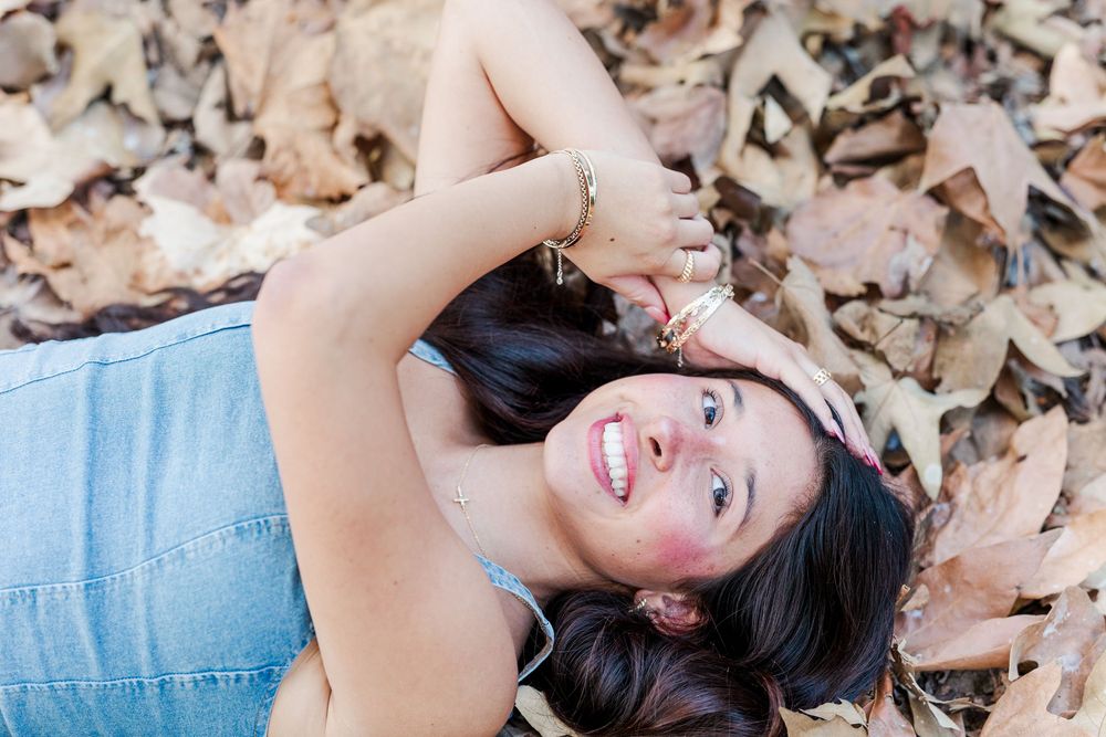 Person playfully poses in fallen autumn leaves wearing blue denim top during outdoor photoshoot.