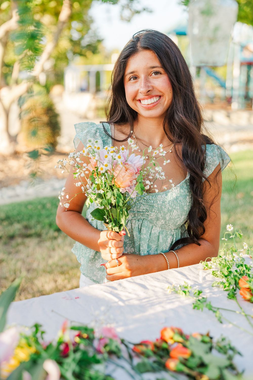 A person in a mint green dress smiles while holding a bouquet of wildflowers outdoors in natural sunlight.