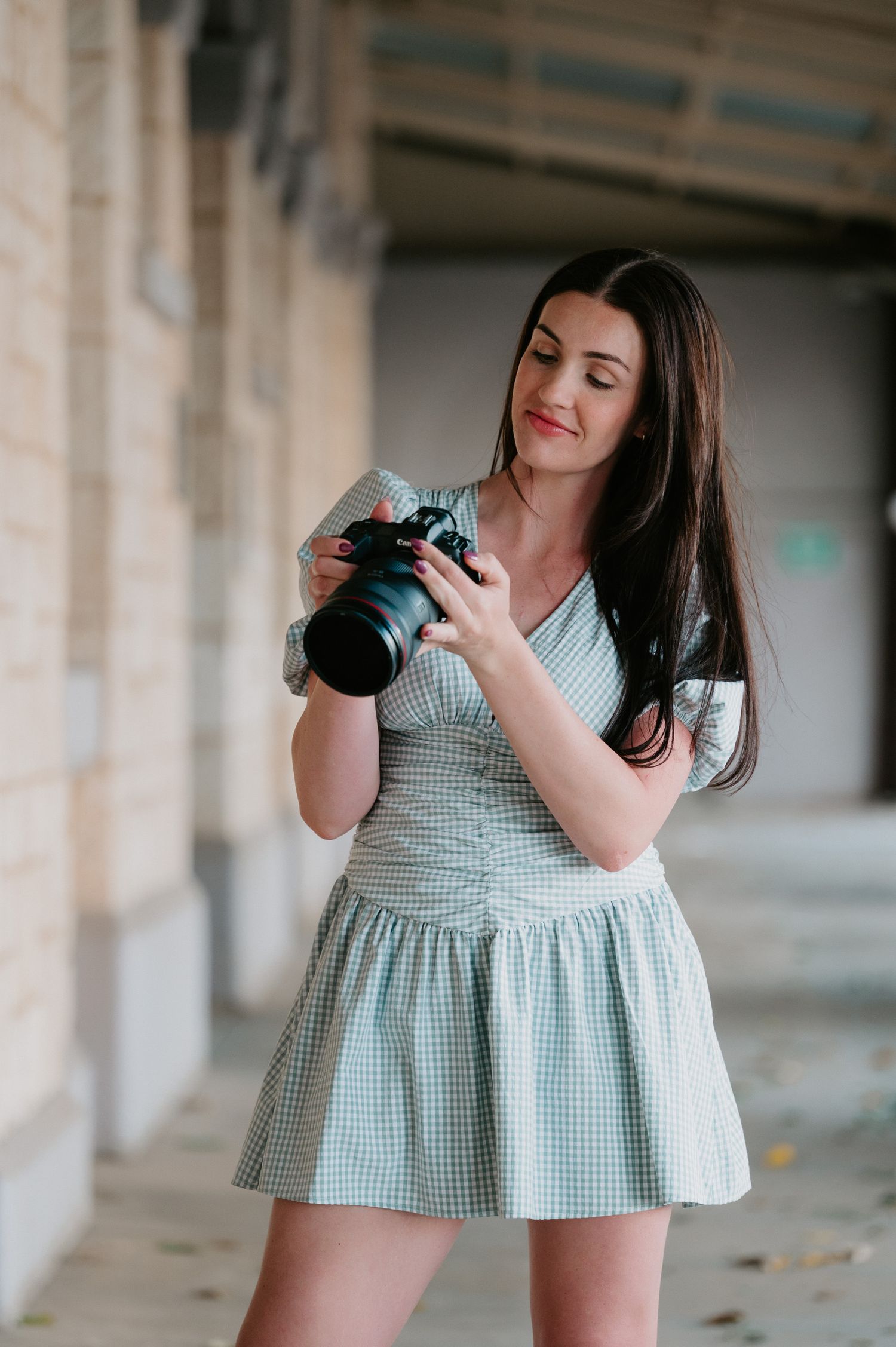 A photographer in a mint gingham dress practices with a camera in an outdoor covered walkway.