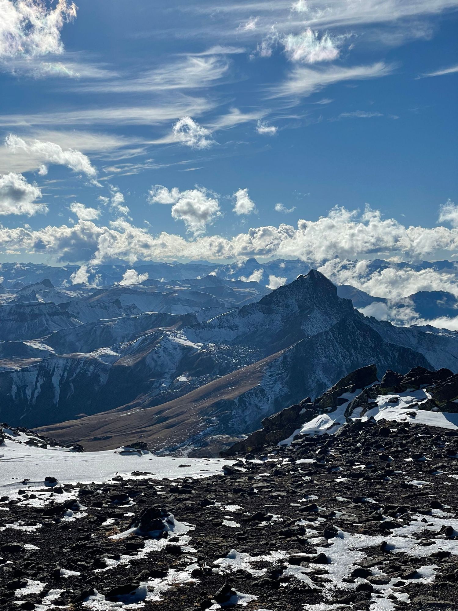 Panoramic mountain landscape with snow-covered peaks and rocky terrain under a dramatic blue sky with white clouds.