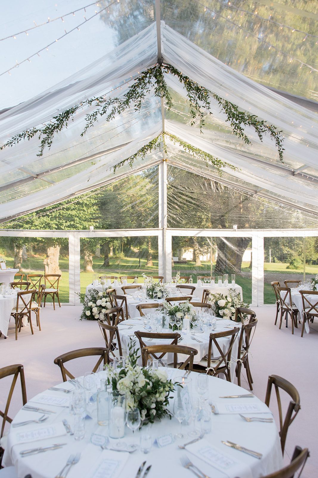 Clear tent wedding reception setup with rustic wooden chairs and elegant white table settings under a transparent canopy.