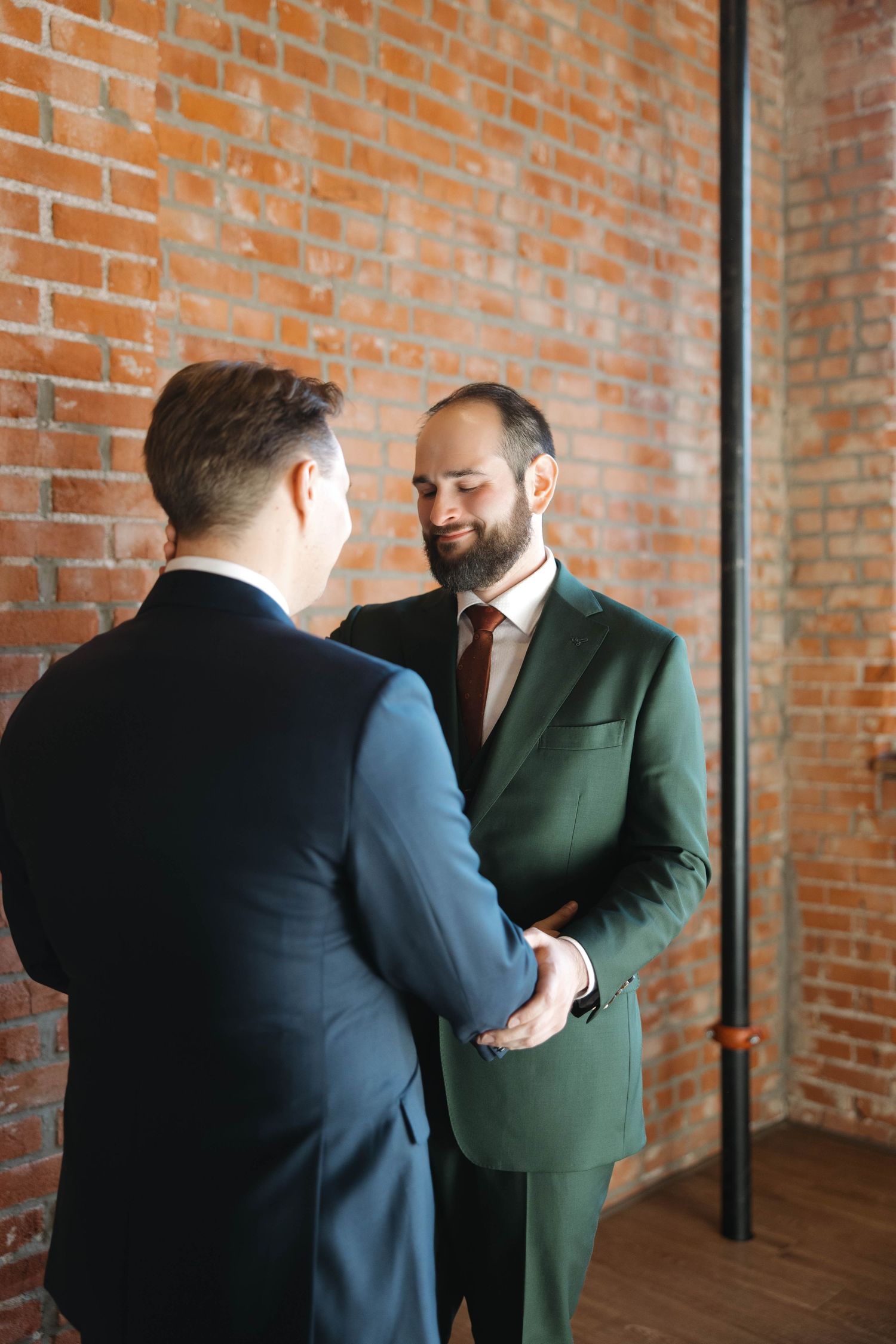 Two men in formal suits shake hands in an industrial-style room with exposed brick walls.
