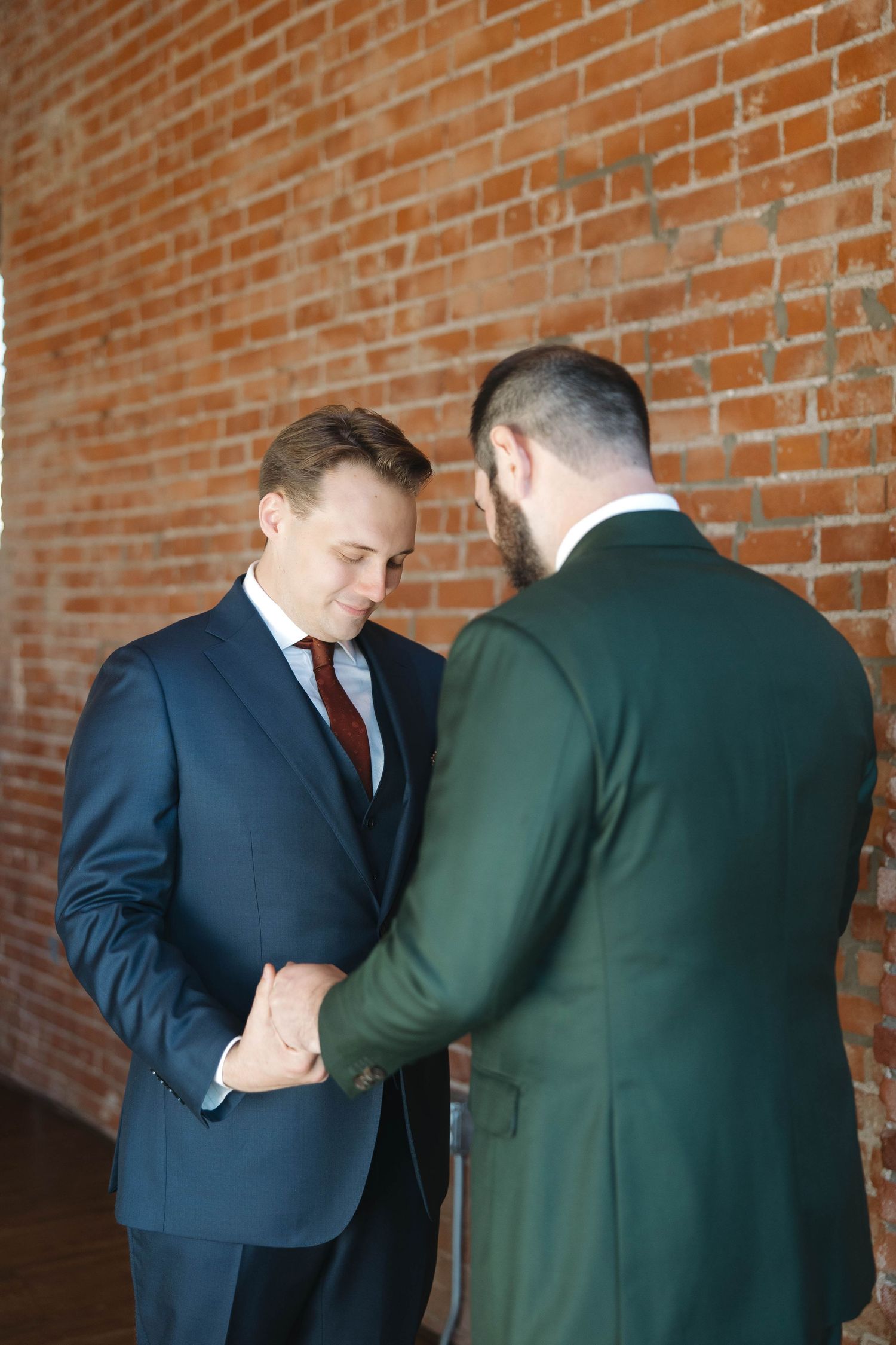 Two people in formal attire share an intimate moment against a rustic brick wall background.