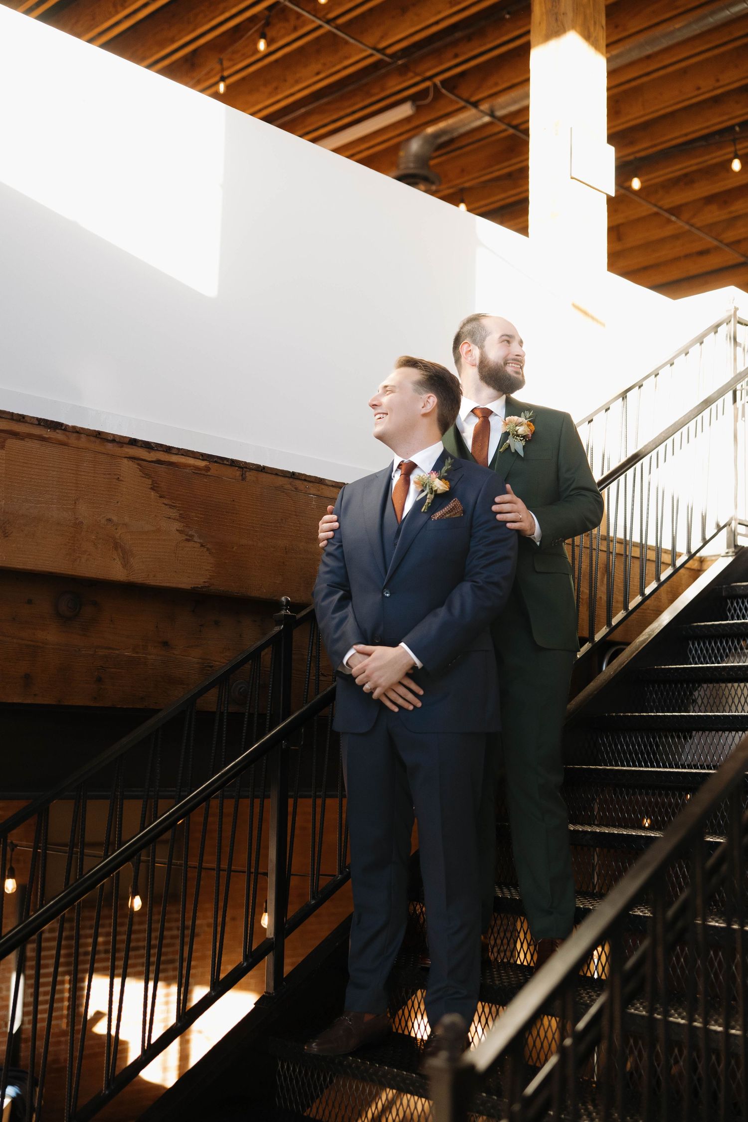 Two people in formal wear standing near black metal stairs in an industrial-style venue.