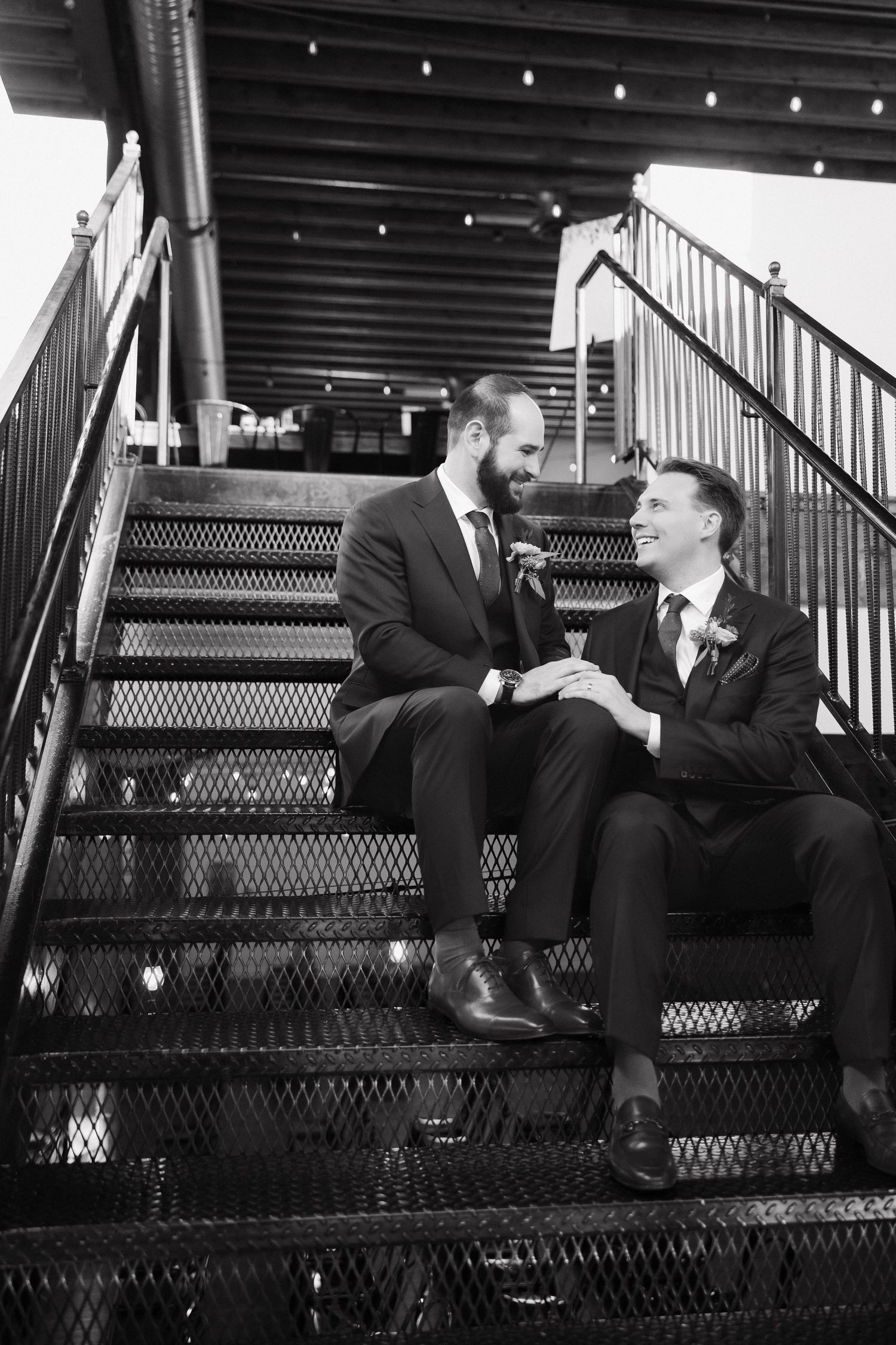 Two people in suits share a candid moment while sitting on stadium bleachers in black and white.