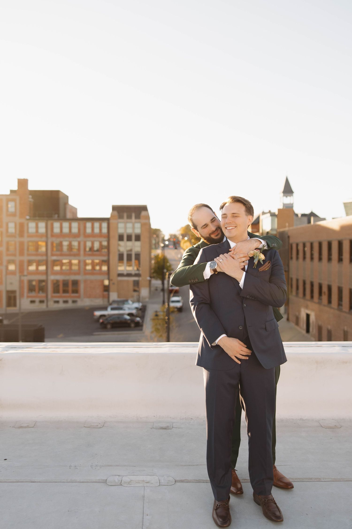 Two people in dark suits share drinks while sitting on a rooftop with urban buildings in background.