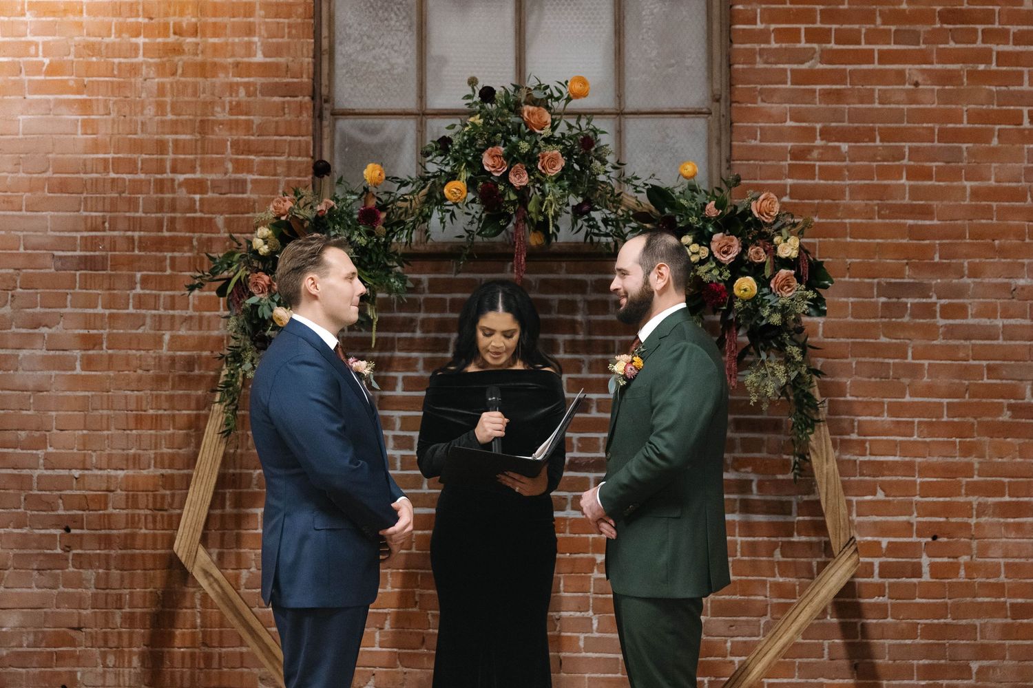 Wedding party gathered in front of exposed brick wall with floral arrangements and wooden decor elements.