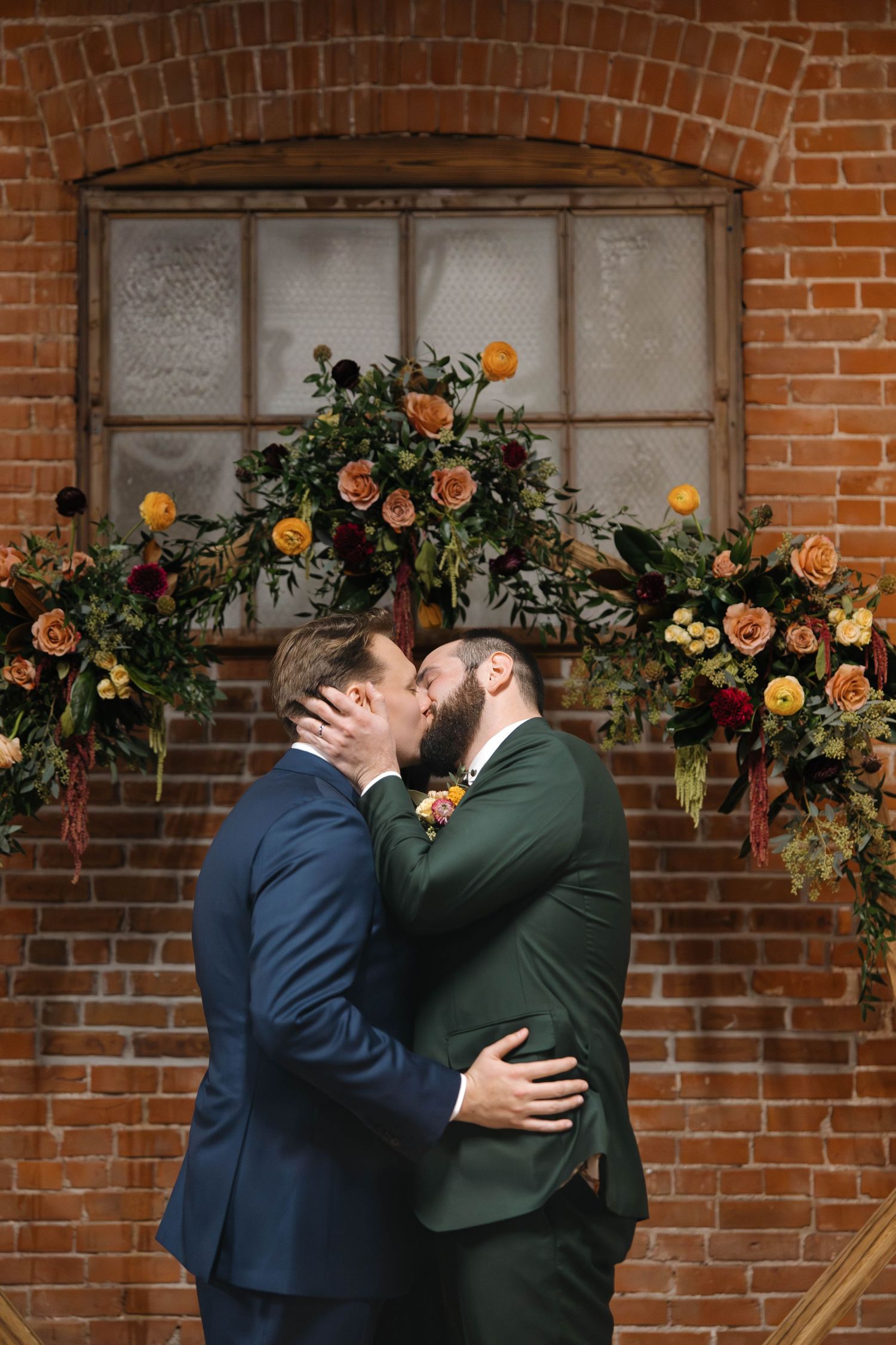 A romantic wedding moment under a floral arch with orange roses against a rustic brick wall and vintage window.