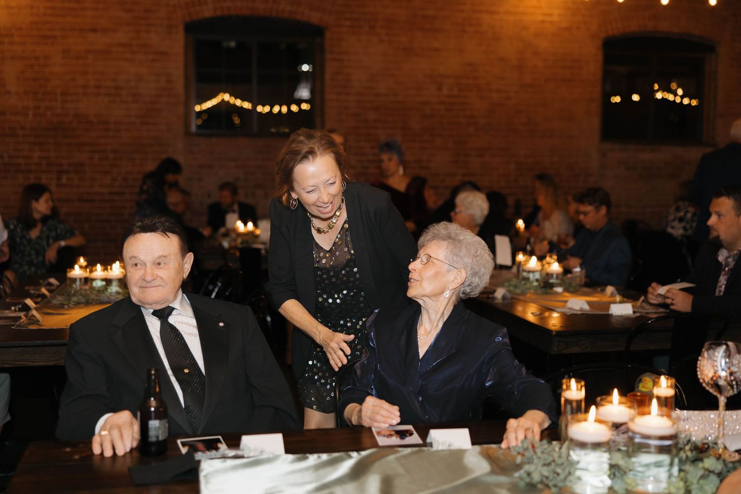Wedding reception guests seated at tables in a dimly lit rustic venue with string lights.