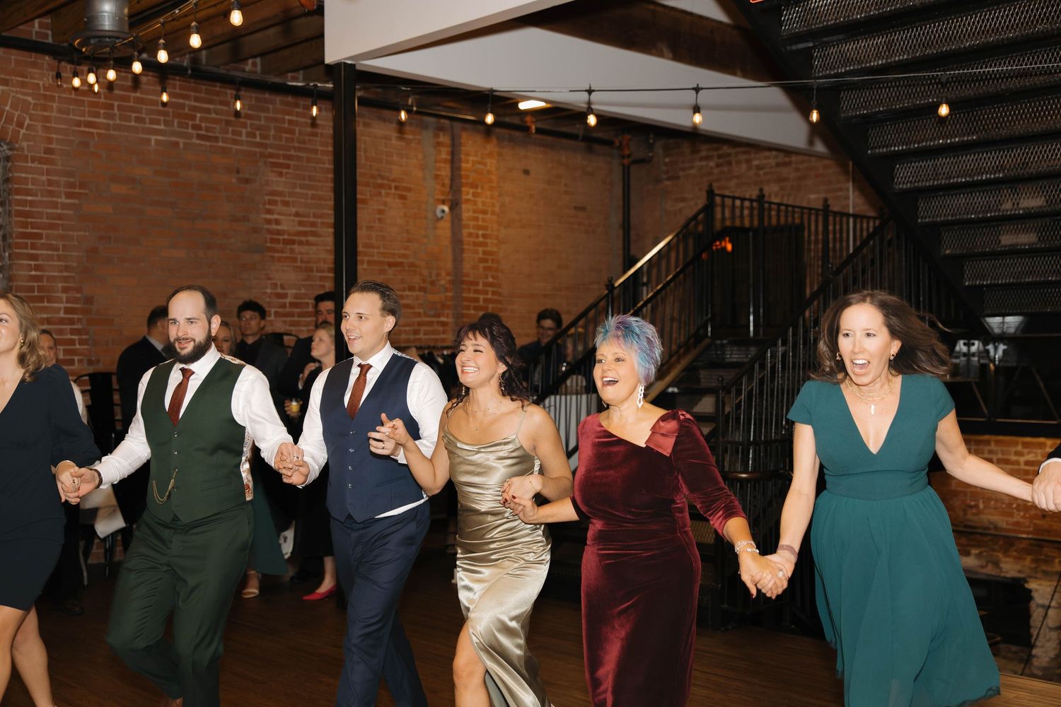 Wedding guests dancing and celebrating in a rustic brick-walled venue with string lights.