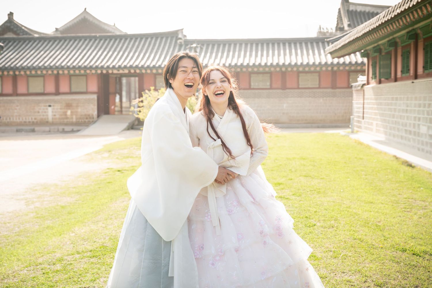 Two people in traditional Korean hanbok clothing walk through a historic palace courtyard on a sunny day.