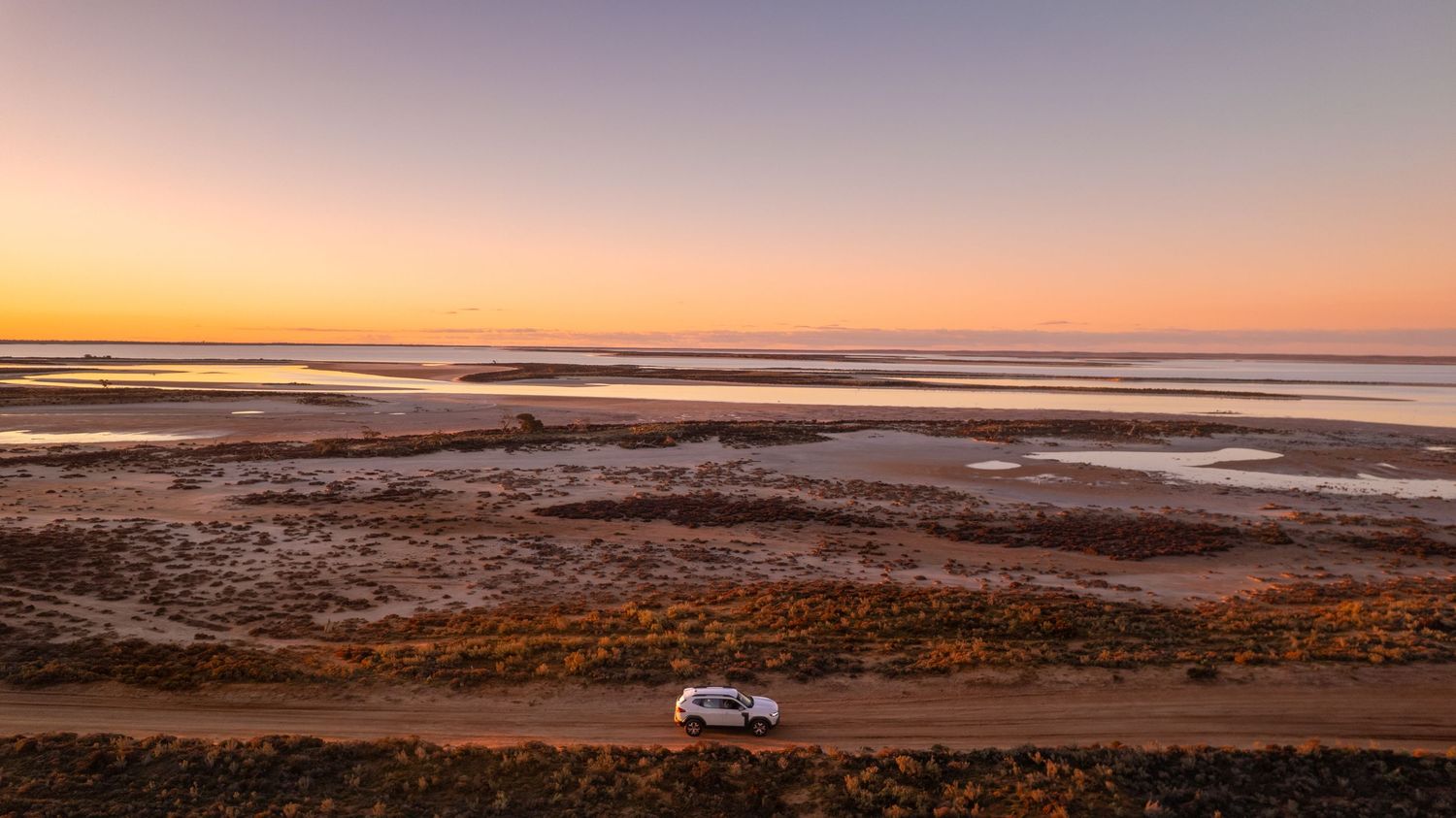 White SUV driving along coastal dirt road at sunset with tidal flats and shallow water pools stretching to the horizon