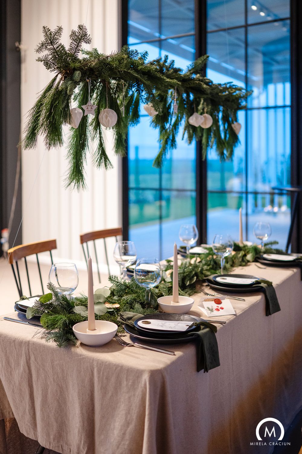 Elegant holiday table setting with hanging evergreen branches and white ornaments above a white tablecloth and wooden chairs.