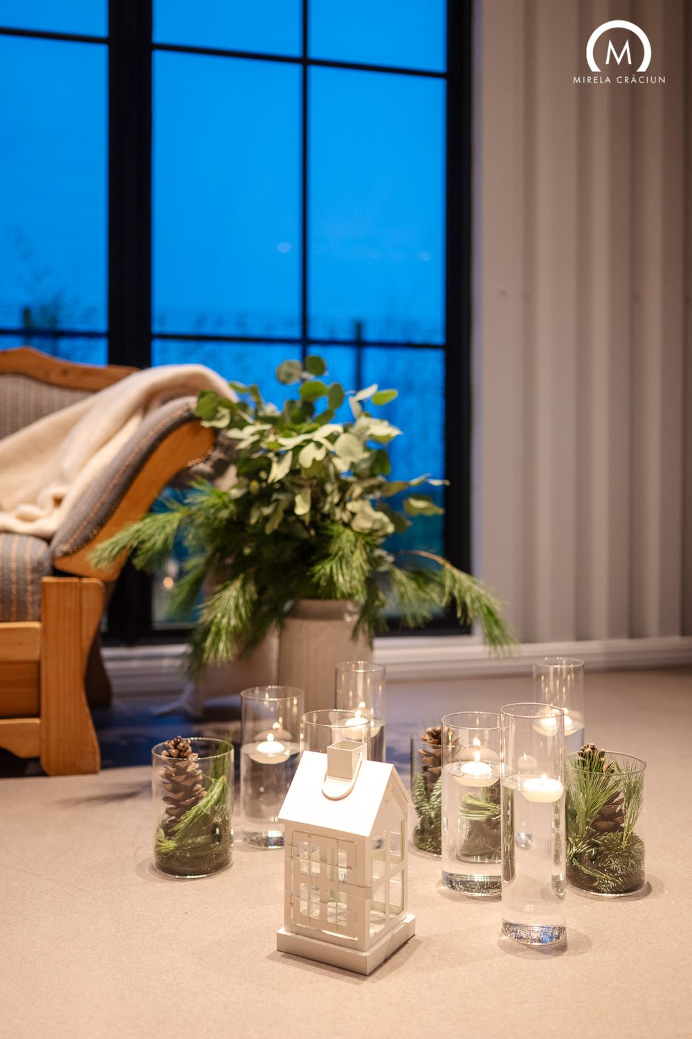 Elegant table decor featuring glass vases with greenery and lit candles against a backdrop of large windows at dusk.