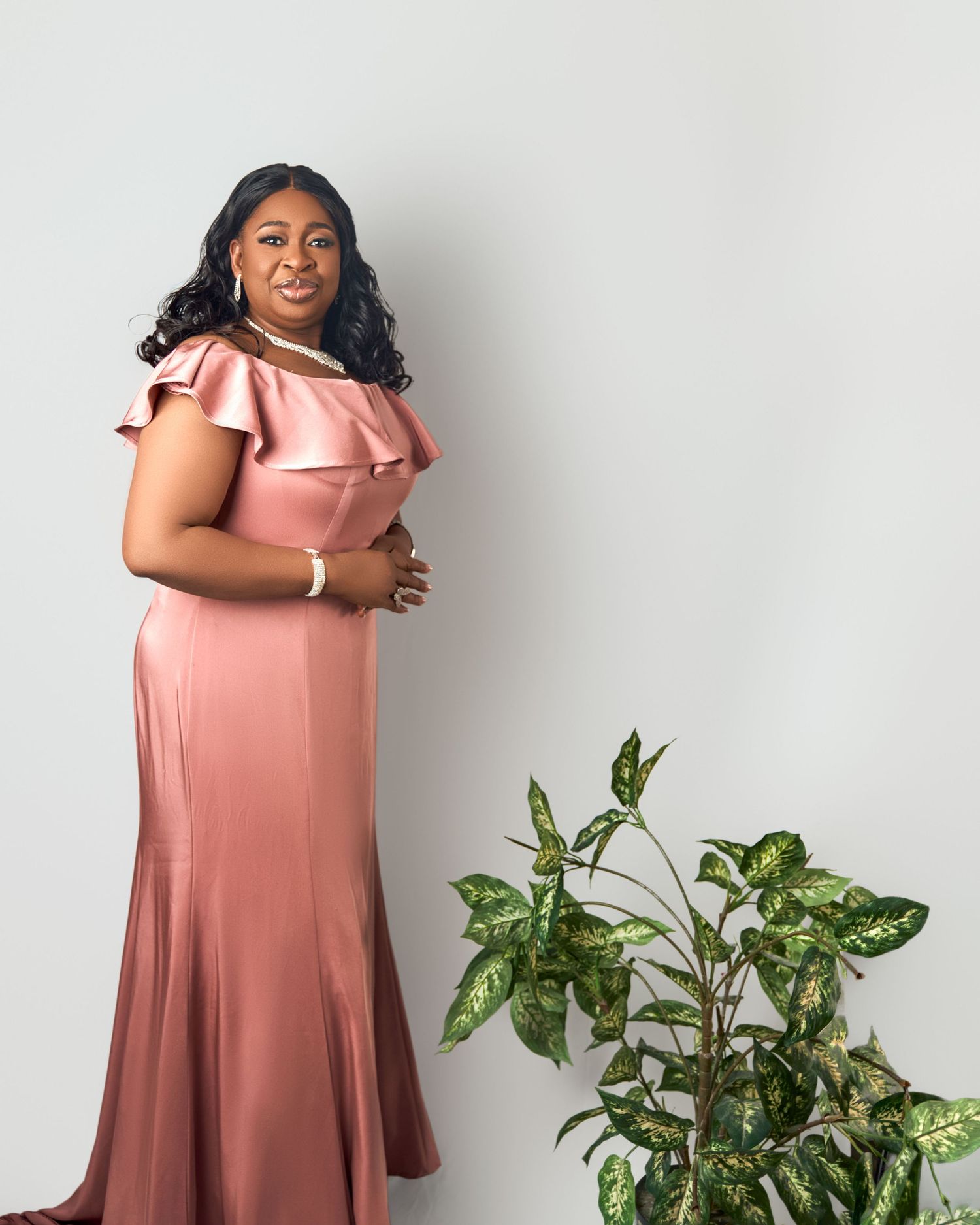 Elegant Black woman poses in rose gold satin off-shoulder mermaid gown beside a green plant.
