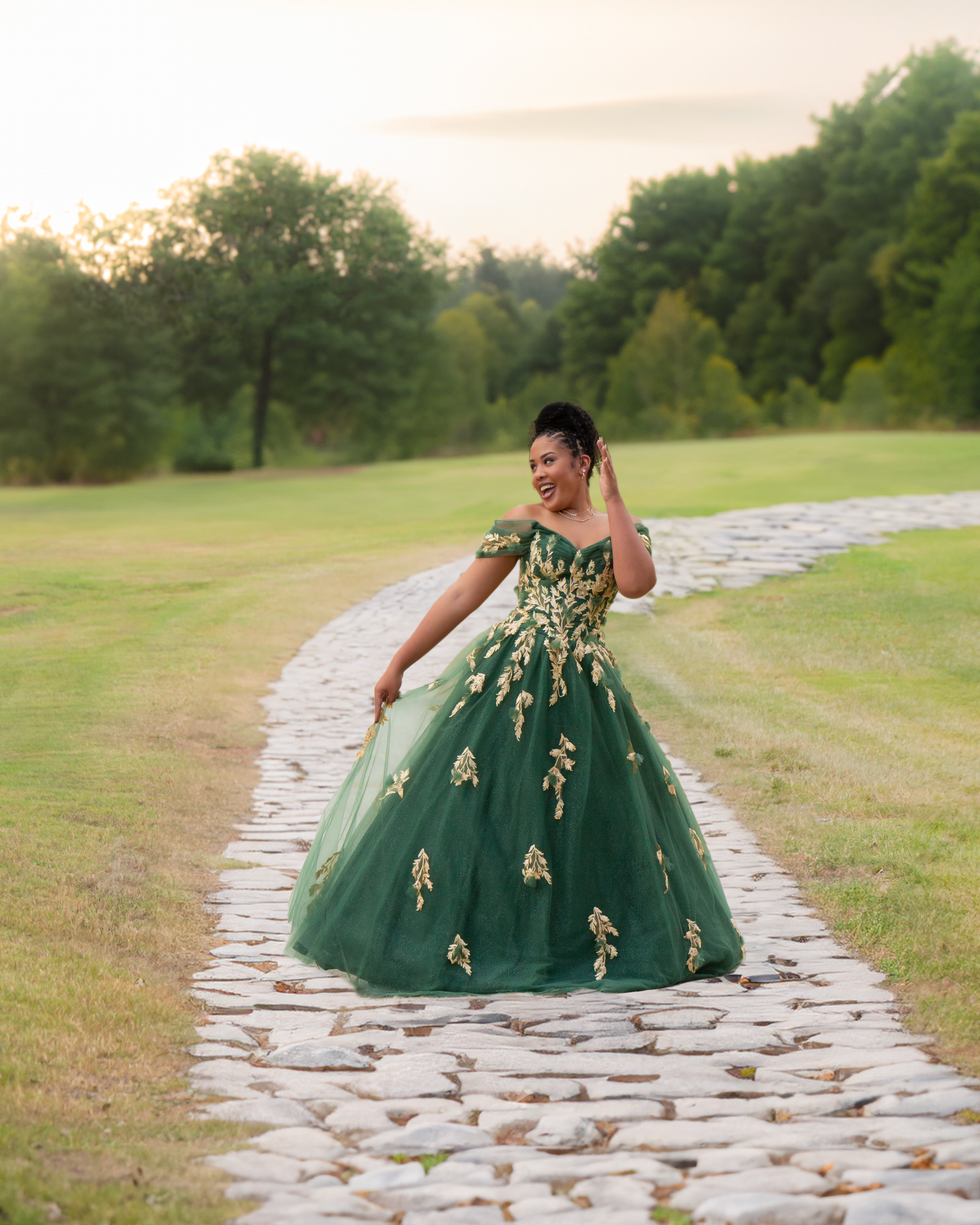 Young woman in emerald green quinceañera ball gown with gold embroidery posing on stone path.