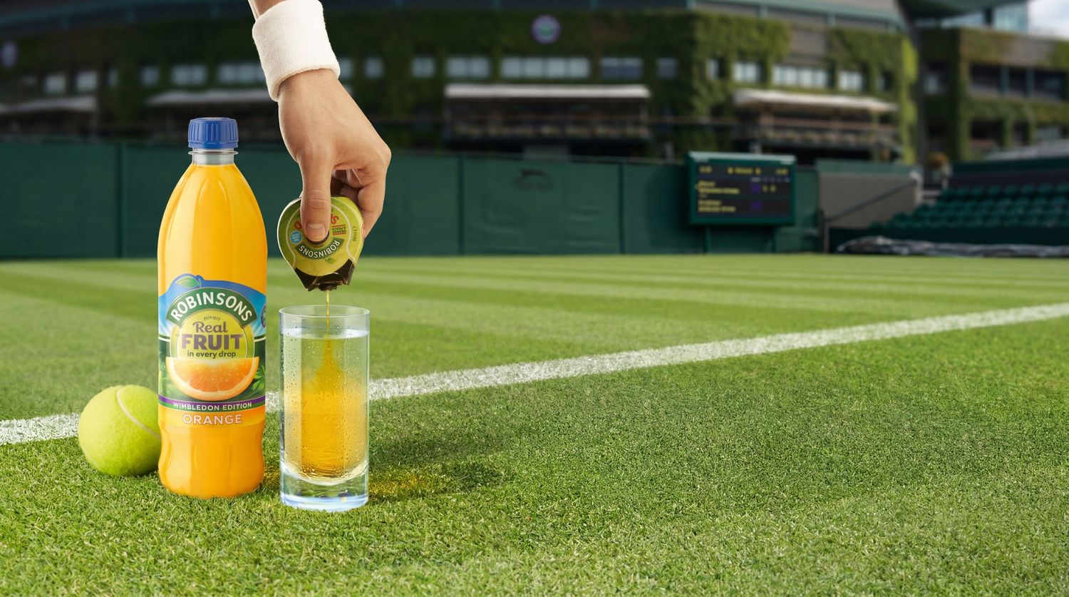 Robinsons Fruit Juice bottle and glass on a Wimbledon tennis court with tennis balls nearby.