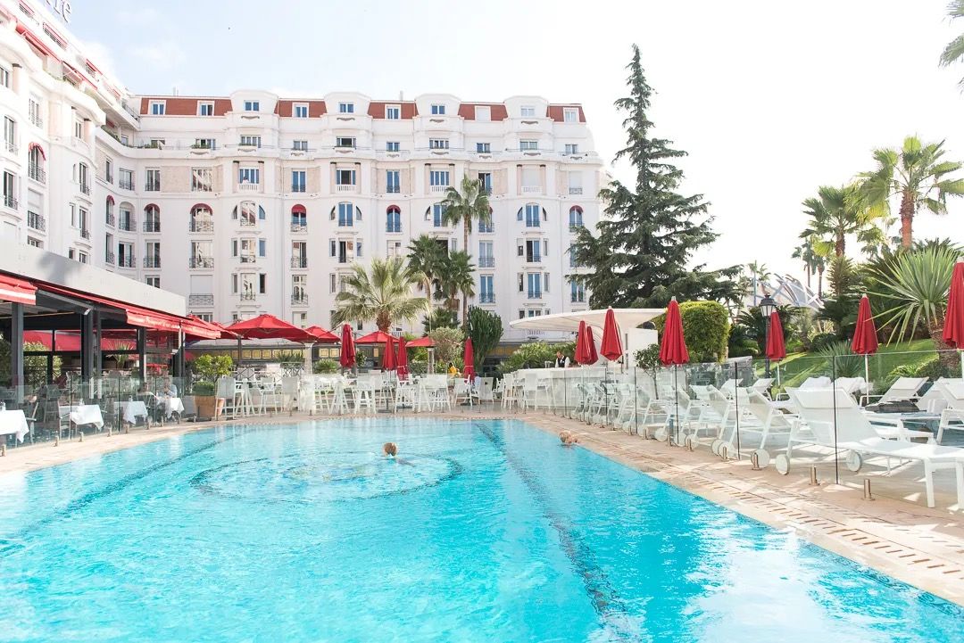 Large hotel with a bright blue outdoor pool surrounded by sun loungers and palm trees.