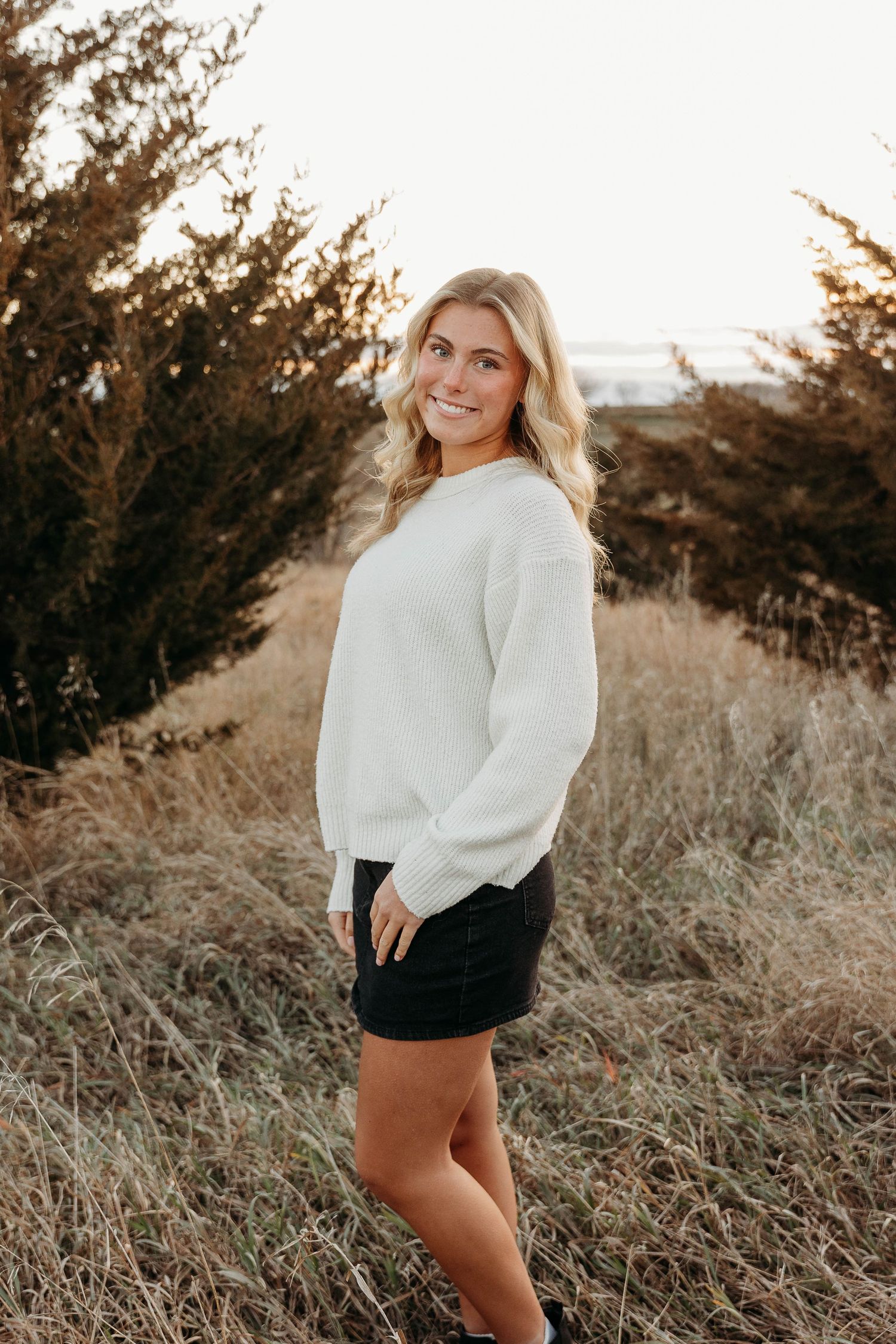 Person in a cozy white sweater and black skirt poses outdoors at golden hour among tall grasses and trees.