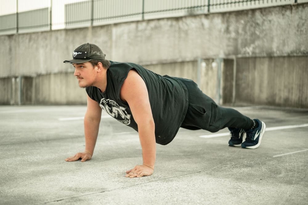 Person in a black tank top and cap performing a push-up exercise on concrete ground outdoors.