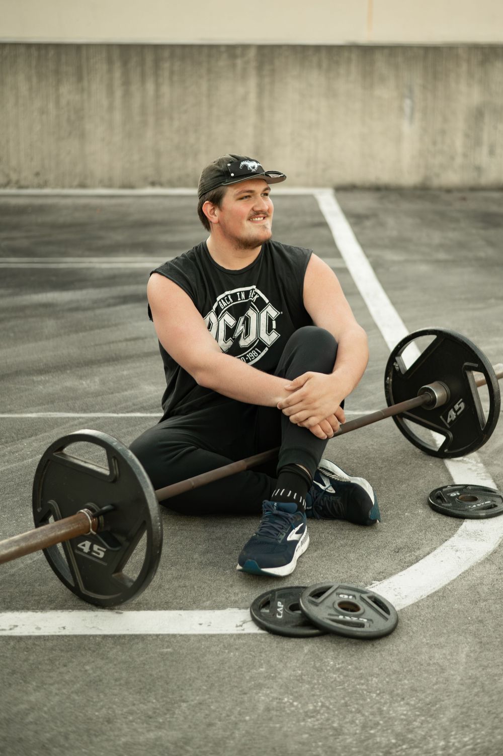 A person in black workout clothes sits on concrete ground with a barbell and weight plates.
