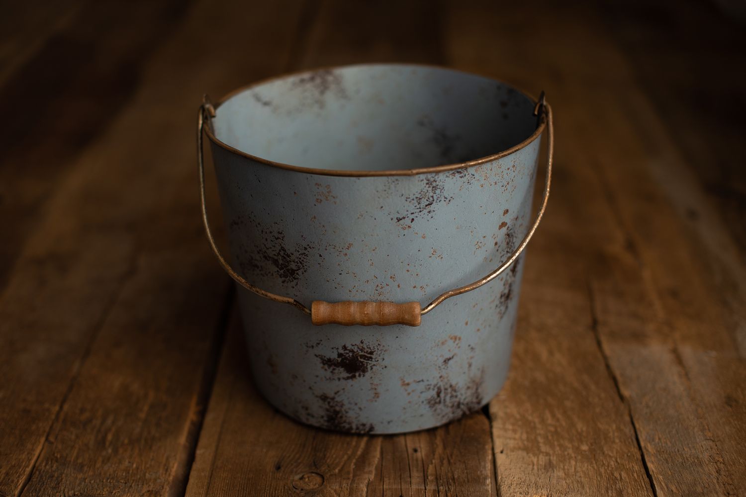 A rustic blue metal bucket with a wooden handle grip, showing aged patina and rust spots on a wooden surface.
