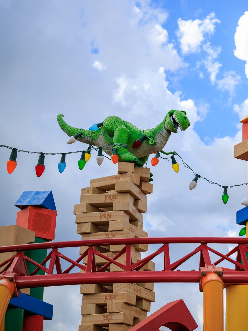 Colorful string lights and toy dinosaurs decorate a wooden block display against a bright blue cloudy sky.