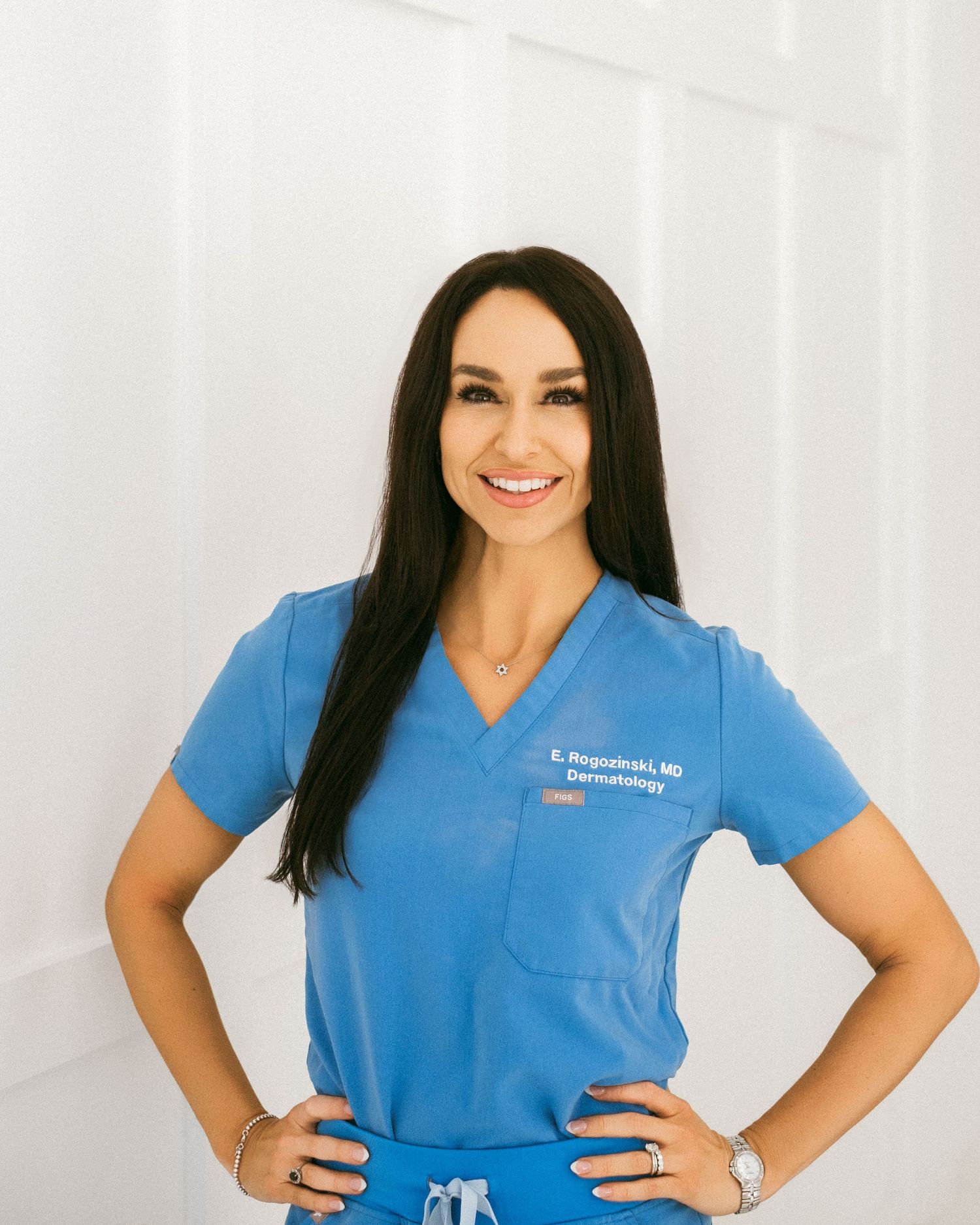 A female dermatologist E. Repolinski MD smiles confidently in blue medical scrubs.