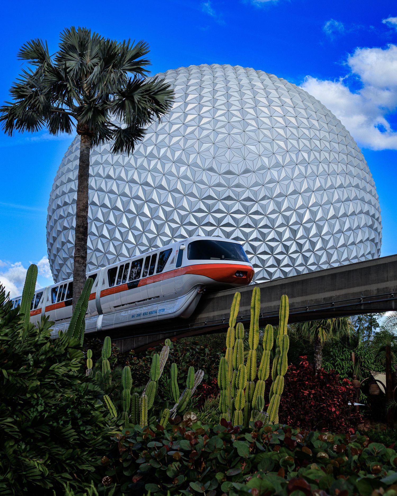 Spaceship Earth geodesic sphere at EPCOT rises against a blue sky with palm trees and desert plants.
