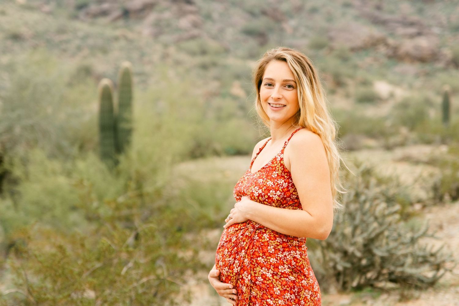 A pregnant woman wearing a red dress and hands on her belly. She's smiling, has blonde hair and has cactus and greenery behind her.