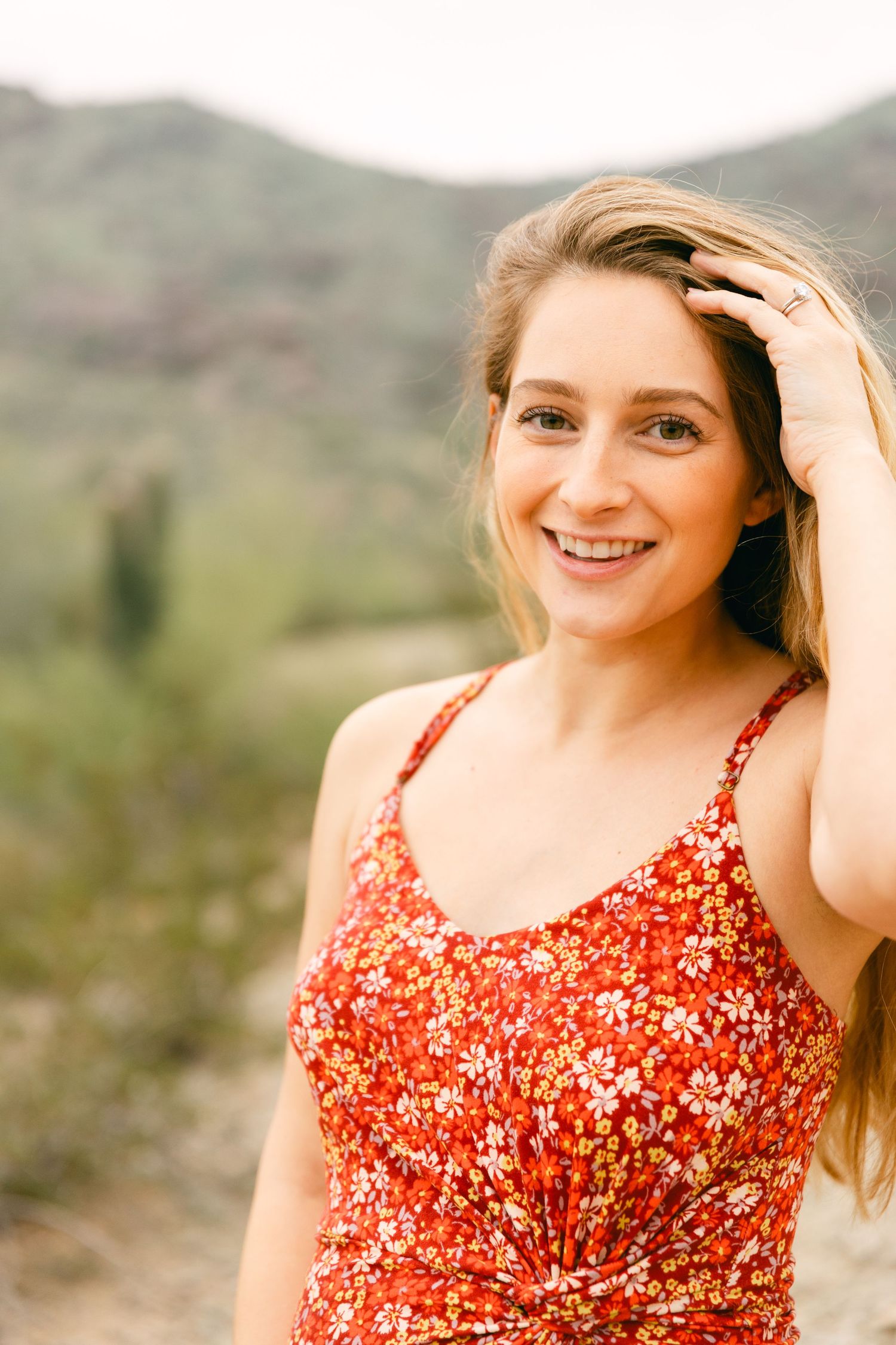 A woman in a red dress is smiling and has a hand in her hair. There's greenery and a mountain behind her.