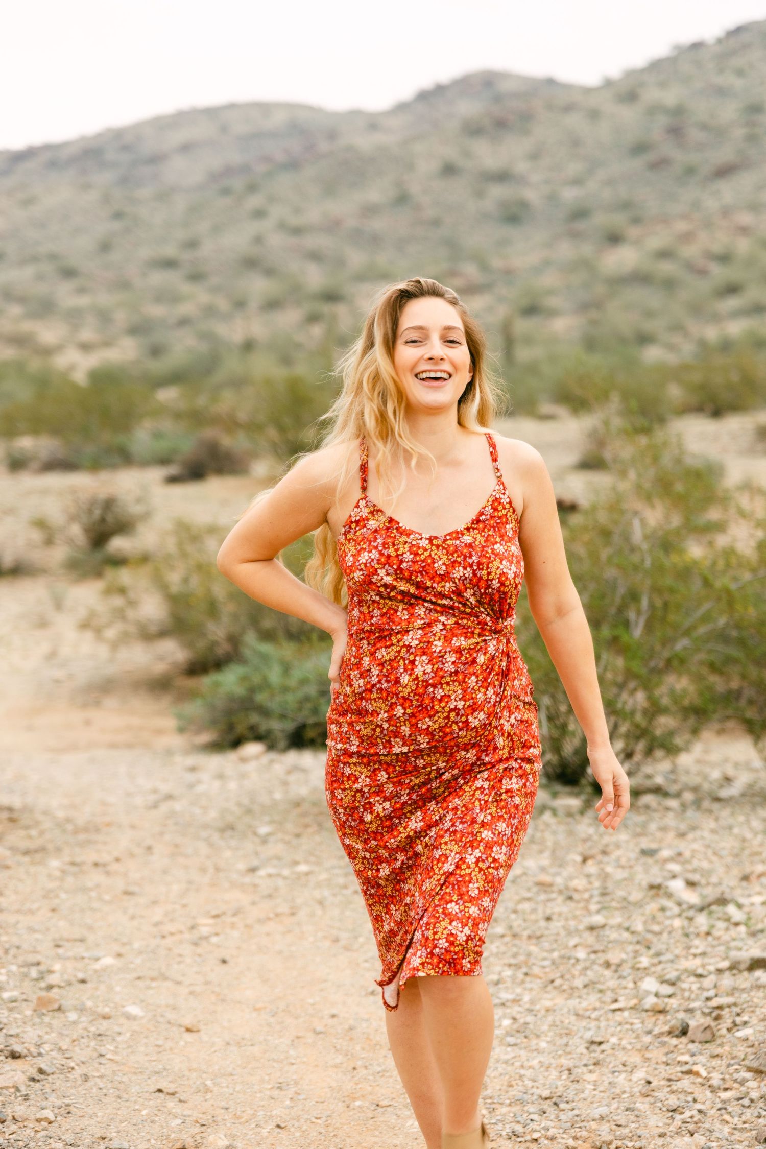 A pregnant woman in a red dress with a hand on her back and is laughing and walking. There's greenery and a mountain behind her.